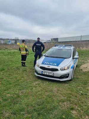 policjant oraz strażak obok radiowozu, stoją tyłem patrząc na nieużytki