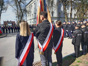 Na zdjęciu stoi tyłem poczet ze sztandarem. Maja na sobie przepasane wstęgi biało-czerwone.