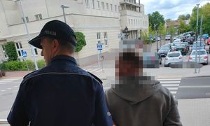 poszukiwany mężczyzna z umundurowanym policjantem przed wejściem do budynku, mężczyzna ma założone kajdanki na ręce