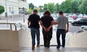 zatrzymany mężczyzna w asyście policjantów, mężczyzna ma założone kajdanki na ręce trzymane z tyłu