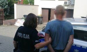 umundurowana policjanta wraz z zatrzymanym mężczyzną na tle budynku i radiowozu, obydwoje stoją tyłem