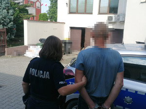 umundurowana policjanta wraz z zatrzymanym mężczyzną na tle budynku i radiowozu, obydwoje stoją tyłem