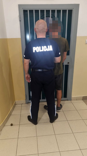 zatrzymany mężczyzna w asyście policjanta na tle pomieszczenia dla osób zatrzymanych