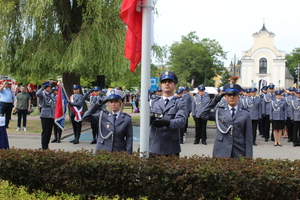 Na zdjęciu stoją policjanci umundurowani, którzy wciągają flagę państwowa  na maszt. W oddali widać stojących na baczność innych umundurowanych policjantów