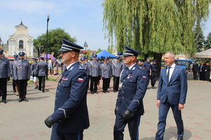 Dwaj umundurowani policjanci idą razem z inna osobą przez plac. w tle widać umundurowanych policjantów.