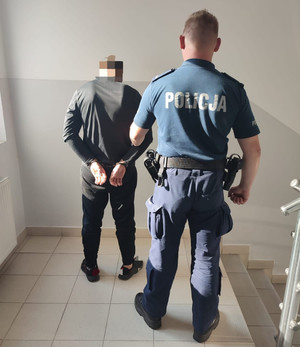 Na zdjęciu umundurowany policjant trzyma pod rękę mężczyznę, który ma założone kajdanki na ręce trzymane z tyłu. Obaj stoją tyłem w pomieszczeniu.