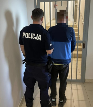 Na zdjęciu stoi umundurowany policjant, który trzyma zatrzymanego mężczyznę pod rękę. Obaj stoją tyłem w pomieszczeniu.