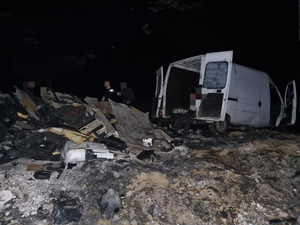 Na zdjęciu jest biały bus z otwartymi tylnymi drzwiami, a przed nim leży wielka sterta odpadów samochodowych.