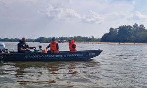 policjanci oraz strażacy siedzący w łódce z napisem Starostwo Powiatowe w Piasecznie, w trakcie patrolu na Wiśle, w tle widać ludzi będących na plaży