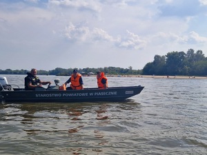 policjanci oraz strażacy siedzący w łódce z napisem Starostwo Powiatowe w Piasecznie, w trakcie patrolu na Wiśle, w tle widać ludzi będących na plaży