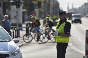 umundurowany policjant wydziału ruchu drogowego stojący przy radiowozie, w tle droga oraz rowerzyści