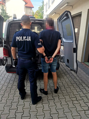 zatrzymany mężczyzna w asyście umundurowanego policjanta, w tle policyjny radiowóz, mężczyzna ma założone kajdanki na ręce trzymane z tyłu