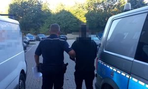 zatrzymany mężczyzna w asyście policjanta, w tle parking policyjny