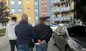 zatrzymany mężczyzna w asyście policjantów, mężczyzna ma założone kajdanki na ręce trzymane z tyłu, w tle bloki mieszkalne