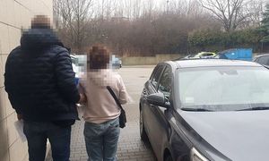 zatrzymana kobieta w asyście nieumundurowanego policjanta, w tle policyjny parking z radiowozami