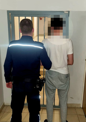 zatrzymany mężczyzna na tle pomieszczenia dla osób zatrzymanych, w asyście umundurowanego policjanta