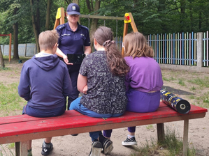 Na terenie placu zabaw siedzi na ławce troje dzieci, które są zwrócone twarzą do umundurowanej policjantki, która trzyma w reku tablet.