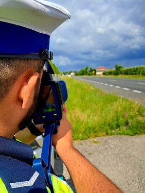 Na zdjęciu jest umundurowany policjant ruchu drogowego, który jest w trakcie czynności pomiaru prędkości. Stoi on na bocznej drodze i urządzenie ma skierowane na główną drogę.