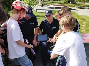 Na zdjęciu siedzą dwaj umundurowani policjanci. Jeden z nich trzyma w ręku tablet . Przed nimi stoi kilkoro chłopców z hulajnogami, którzy oglądają filmik na tablecie, który trzyma policjant.