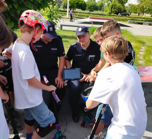 Na zdjęciu siedzą dwaj umundurowani policjanci. Jeden z nich trzyma w ręku tablet . Przed nimi stoi kilkoro chłopców z hulajnogami, którzy oglądają filmik na tablecie, który trzyma policjant.