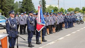 Na zdjęciu stoją w szyku umundurowani policjanci. Na początku rzędu stoi dowódca uroczystości a za nim poczet flagowy i inni policjanci.