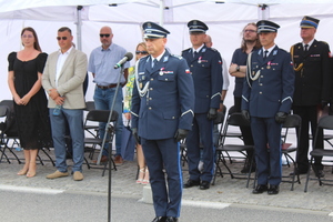 Przed mikrofonem stoi na baczność umundurowany policjant, a za nim pod namiotem stoją osoby cywilne oraz umundurowani policjanci.