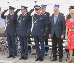 Pod namiotem stoi trzech umundurowanych policjantów, którzy oddają honor, obok nich są osoby ubrane po cywilnemu.