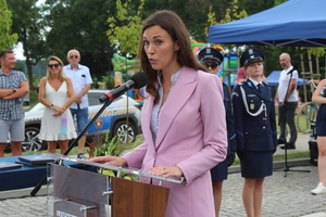 Przy mównicy stoi kobieta ubrana na różowo. W tle widać innych policjantów oraz osoby cywilne.