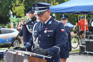 Przy mównicy stoi umundurowany policjant. W tle widać innych funkcjonariuszy.