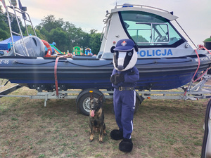 Na tle łodzi rzecznej Policji stoi policyjna maskotka borsuk, obok niej siedzi policyjny pies.