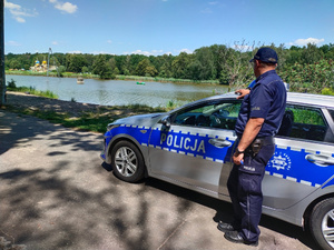 Umundurowany policjant stoi przy radiowozie. W tle widać wodę i basen.