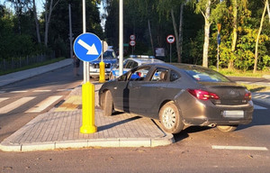 Ciemny samochód osobowy stoi na wysepce drogowej pomiędzy znakami drogowymi . Tył auta jest częściowo na ulicy. w tle widać dwa radiowozy oraz terem leśny.