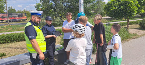 Na zdjęciu stoi umundurowany policjant ruchu drogowego obok umundurowanej policjantki z prewencji, która w ręku trzyma tablet. Przed funkcjonariuszami stoją dzieci, niektóre maja kask na głowie i oglądają filmik na tablecie, który trzyma policjantka. Wszyscy są na skejt parku.