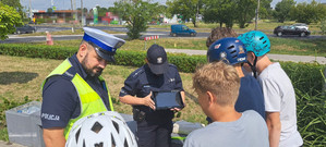 Na zdjęciu stoi umundurowany policjant ruchu drogowego obok umundurowanej policjantki z prewencji, która w ręku trzyma tablet. Przed funkcjonariuszami stoją dzieci, niektóre maja kask na głowie i oglądają filmik na tablecie, który trzyma policjantka. Wszyscy są na skejt parku.