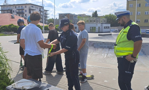 Na zdjęciu jest umundurowany policjant z ruchu drogowego oraz umundurowana policjantka z prewencji, która zakłada odblaskowa opaskę jednemu ze stojących obok niej chłopaków. Wszyscy są na skejt parku.