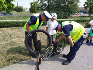 Policjanci z ruchu drogowego sprawdzają oznaczenie roweru, który stoi do góry kołami. Obok nich stoi chłopiec, który ma kask na głowie.