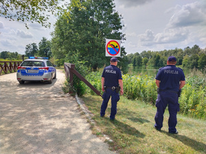 Dwóch umundurowanych policjantów patrzy na wodę. Policjanci stoją obok znaku &quot;zakaz kąpieli&quot;, w tle widać radiowóz i zarośla przy wodzie.