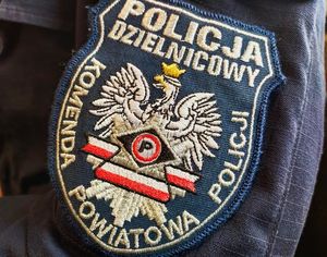 Na ramieniu policyjnego munduru jest policyjna plakietka. Jest na niej napis: Policja, Dzielnicowy, Komenda Powiatowa Policji. Na środku plakietki jest orzeł biały w koronie.