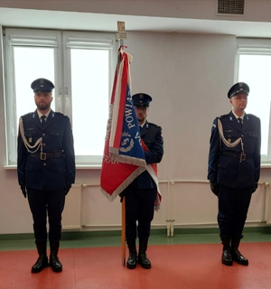 Na zdjęciu stoi na baczność trzech umundurowanych policjantów. Środkowy policjant trzyma w ręku sztandar Policji.