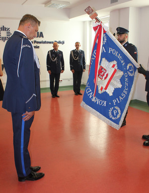 Na zdjęciu są umundurowani policjanci. Dwaj stoją na baczność pod ścianą. Z przodu stoi policjant który ma opuszczoną głowę przed sztandarem, na którym jest napis Komenda Powiatowa Policji w Piasecznie.