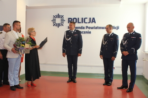 Na zdjęciu jest trzech umundurowanych policjantów. Obok nich stoją osoby ubrane po cywilnemu.Kobieta w reku trzyma otwarta teczkę, obok niej stoi mężczyzna z kwiatami.