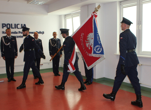 Trzej policjanci maszerują, jeden z nich niesie na ramieniu sztandar komendy. Pod ścianą stoi trzech policjantów na baczność.