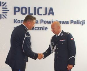 dwaj umundurowani policjanci witają się. Na tle napisu Policja, Komenda Powiatowa Policji w Piasecznie.