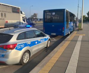 oznakowany radiowó9z stoi w zatoczce autobusowej. Przed nim jest niebieski autobus, do którego wsiada osoba. Obok zatoczki autobusowej jest ulica, po której jedzie inne auto.