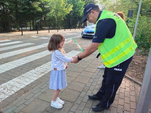 Umundurowany policjant w kamizelce odblaskowej zakł9ada na rękę dziewczynki pasek odblaskowy.