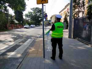 Umundurowany policjant ruchu drogowego stoi przy przejściu dla pieszych. w ręku trzyma paski odblaskowe i jest twarzą zwrócony w kierunku drogi.