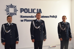 Trzej umundurowani policjanci stoją w szeregu W tle na ścianie widać logo i napis Policja, Komenda Powiatowa Policji w Piasecznie.