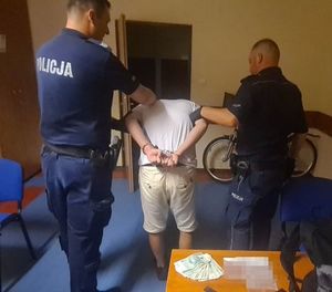 Dwaj umundurowani policjanci trzymają zatrzymanego mężczyznę pod ręce. Mężczyzna ma założone kajdanki na ręce trzymane z tyłu. Wszyscy stoją tyłem. Obok jednego z policjantów stoi krzesło. Za wszystkimi są otwarte drzwi obok których stoi rower. Na zdjęciu przed policjantem jest stolik, na którym leżą rozłożone pieniądze.