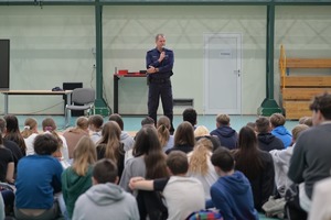 policjant w trakcie spotkania z młodzieżą. Uczniowie siedzą na hali sportowej, funkcjonariusz z mikrofonem stoi przed nimi