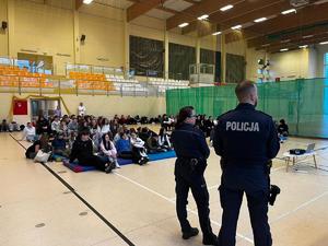 policjanci, w tle siedzący na hali sportowej uczniowie szkoły